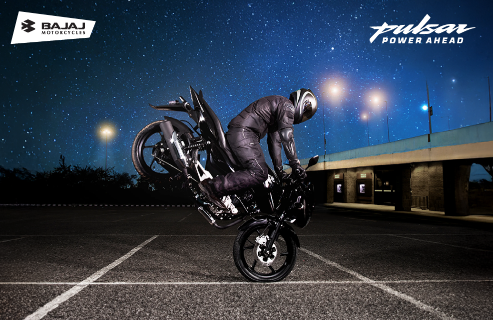 A man performs a motorcycle trick on a Bajaj Pulsar 150, showcasing skill and balance in an open space.