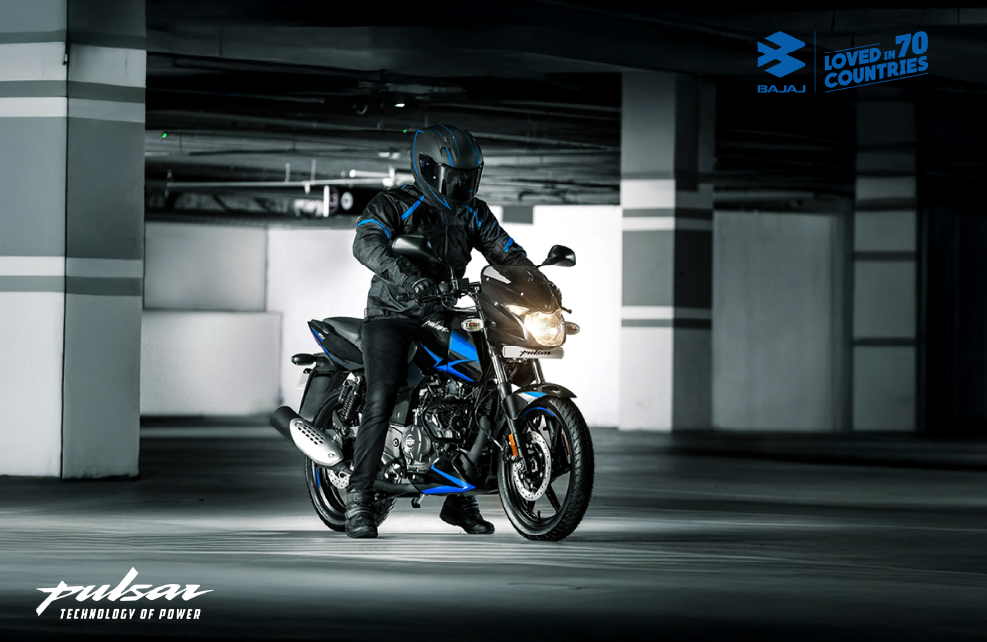 In a dark parking garage, a man is seen on a black and blue Bajaj Pulsar 150 motorcycle.