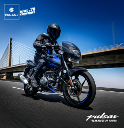 A man is riding a black and blue Bajaj Pulsar 150 motorcycle on the road, with a bridge visible in the background.